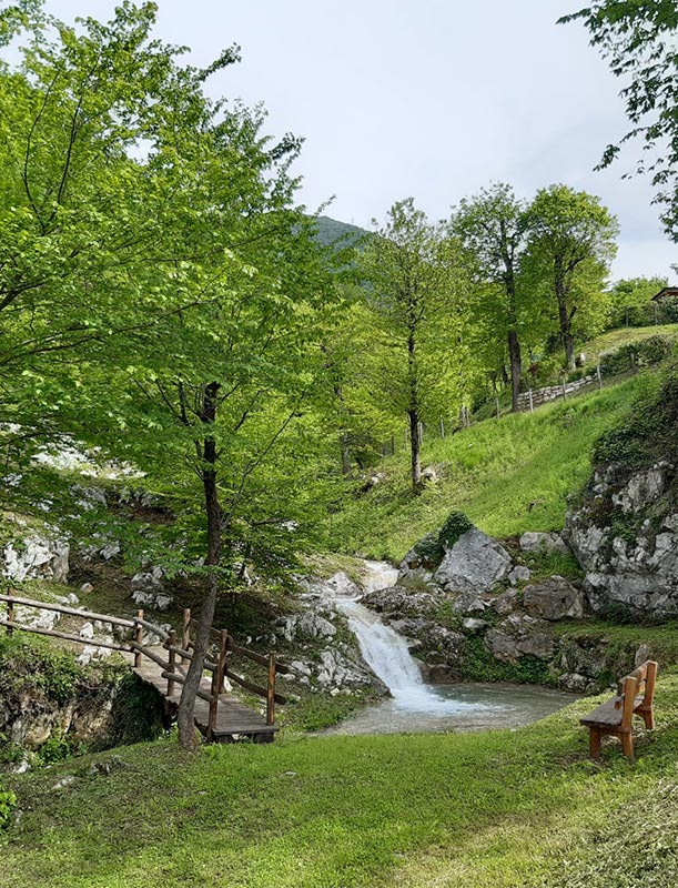 Cascata tra le rocce con passerella in legno e panchina nel verde castagneto del Piccola Valle – Valley Resort